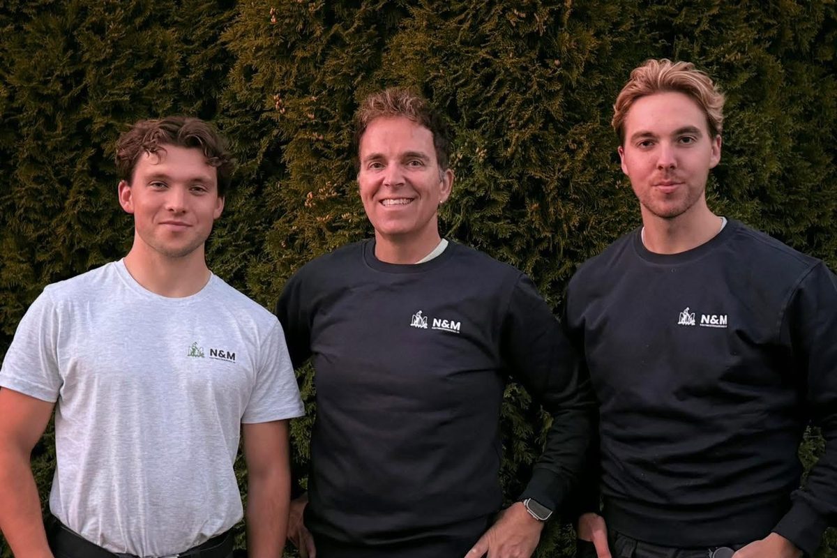 Tre fra N&M Vaktmesterservice i arbeidsklær med logo, foran en hekk